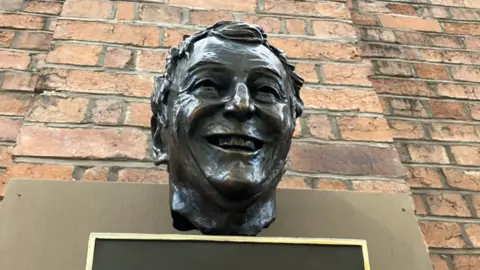 A bronze bust of a man's head with short hair in front of a brick wall