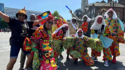 Toby Friedner/BBC A group of people wearing colourful outfits of red, yellow, and green pose for the camera. Many have their arms out in celebration as they smile. It is a sunny day.