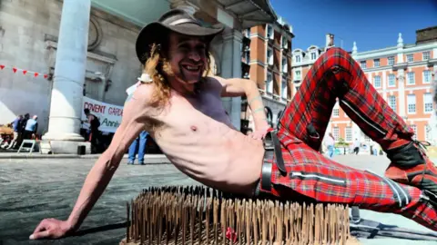 A man lying on a bed of nails. He has no top on but is wearing a cowboy hat and tartan punk-style trousers. He is smiling and has long hair.