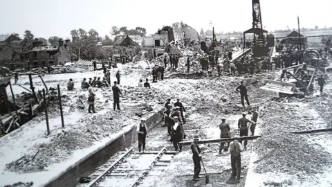 Soham Museum Workmen clearing debris-strewn Soham railway line with demolished buildings in the background, 1944