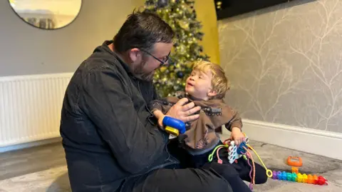 BBC John wears glasses and has a beard. He is sitting on the living room floor with his son sitting on his knee. His son has blonde hair and is wearing a brown sweatshirt. He is holding toys in both his hands.