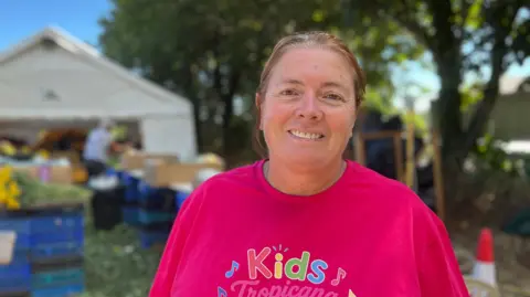 Marina Noel pictured smiling outside the Grouville battle shed. There are boxes of flowers visible in the background. She has dark hair in a pony tail, and is wearing a pink tshirt.