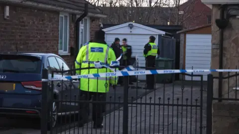 Adam Haggy Hargreaves Four police officers wearing black uniforms and high visibility jackets stand on a driveway between two houses. The officers are standing behind a black metal gate which is partially covered with blue and white police tape.