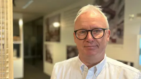 Steve McIntyre has short white hair and wears dark rimmed glasses. He is wearing a white shirt. He is pictured in his office, standing beside a 3D scale model of a skyscraper. 