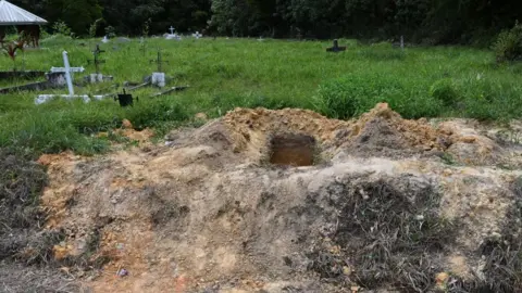An open grave at the site in Cumuto, the rest of which shows graves with crosses on overgrown grass