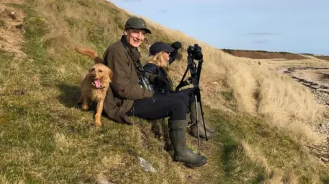 Adrian Crellin Prof Crellin sat on a hillside with his wife and dog with a camera on a tripod in front of him 