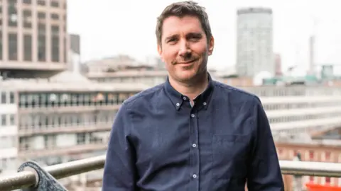 Jonathan Patric Gilchrist wearing a navy blue shirt and smiling. He has brown slightly spiky hair and is standing on a balcony with a cityscape behind him that is blurred out.