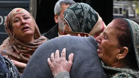 A group of people mourn the death of their relatives following a suicide bombing at a mosque in Islamabad. The woman are wearing head scarves and look distraught. One woman is hugging someone in a grey jumper, their head is obscured by her embrace.