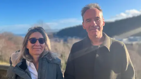 Prof McStay and Prof Bakir are standing outside and both looking at the camera with hillsides behind them. Prof Bakir has shoulder-length hair and sunglasses. She is wearing a grey winter coat. Prof McStay has short grey hair and is wearing a green wool coat.