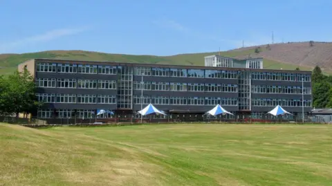 An old school building across four storeys set next to a playing field and with green rolling hills in the background