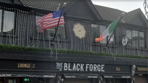 The sign of a pub that says The Black Forge in white writing on a black background. An American flag and an Irish tricolour are above. 
