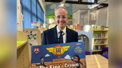 A smiling Rob Johnson with one of his books from the series Let's Go See, called The King's Crown, at the school.