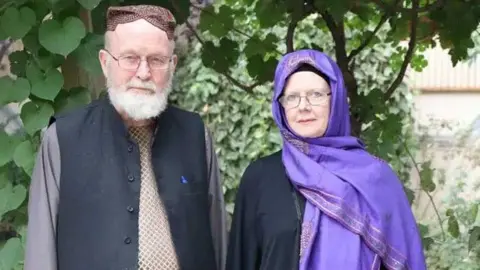 A man, with white beard and glasses, wearing a dark vest stands next to a woman wearing a black top and purple head scarf and glasses, in front of greenery.