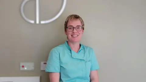 Rachele Crozier Shows a woman with short fair hair and glasses smiling. she is wearing a jade-coloured dental outfit and is standing in front of a wall.