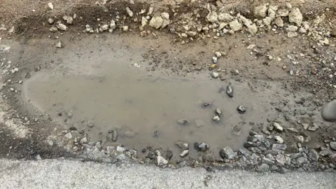 One of the potholes which appears quite deep, with a puddle inside and dirt and rocks surrounding it