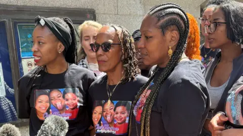 PA Media TJ Jacobs, Vyleen Maximen and another family member address the media outside court. They are wearing T-shirts with Cher's face on it. Supporters are gathered behind them.
