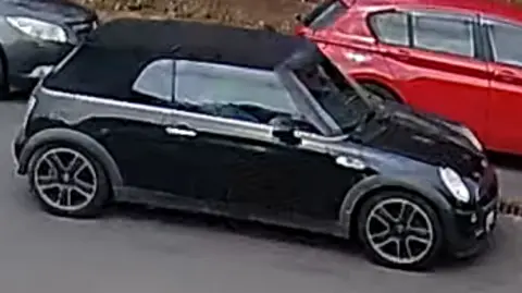 A black Mini Cooper is seen from the side as it drives along a minor road