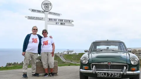 Dave Whittington A couple standing under the Land's End finger post and next to a classing car in British Racing Green