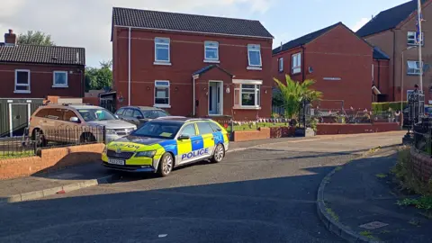A PSNI car is parked outside a house. The car is yellow and blue and states police on the front and side. 