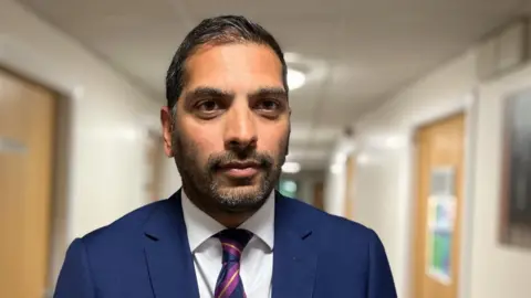 Steve Russell is in a navy suit standing in a hospital. He is wearing a striped purple, yellow and navy tie.