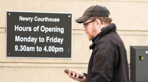 Pacemaker Chris Fulton is pictured holding his telephone  and wearing a achromatic  cap. He is wearing achromatic  framed glasses and a achromatic  coat. He is walking past   a motion   that says Newry Courthouse, Hours of opening, Monday to Friday, 9.30am to 4.00pm.