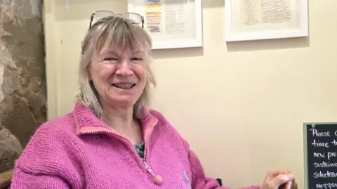 Paul Moseley/BBC Jayne Rounce is smiling at the camera, with her glasses propped up on top of her head. She is wearing a bright pink woolly fleece-type top.