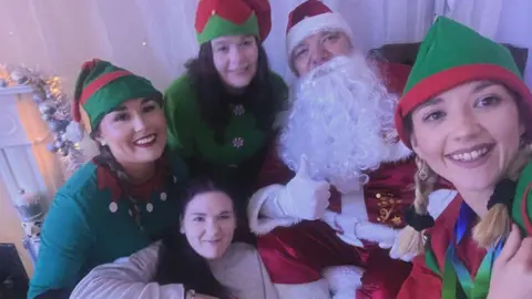 A group of women dressed as Christmas elves pose with a man dressed as Father Christmas. Santa has a bush white beard, red jacket with white fur trim and a red and white hat. He is doing a thumbs up to the camera with a white gloved hand. The elves are wearing green tops and green and red pointed hats.