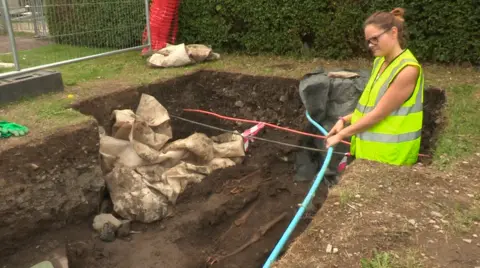 A woman with brown hair tied up wearing a high‑visibility vest stands beside a rectangular excavation trench outdoors, holding a cable or hose. Exposed soil, stones, protective netting and pipes are visible inside the trench, with fencing and grass in the background.
