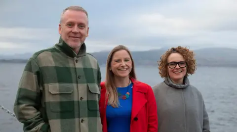 James Chapelard A man and two women stand with their backs to the River Clyde, smiling at the camera. The man has blonde hair and a beard and is wearing a green and beige checked jacket. The woman to his left has long blonde hair, a bright red trench coat and a royal blue top. The woman to her left has short brown curly hair, dark-rimmed glasses and a grey fleece.