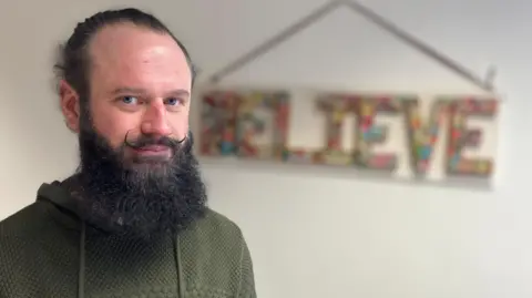 Jon Wright/BBC A man with a beard stands in front of a colourful sign which says Believe