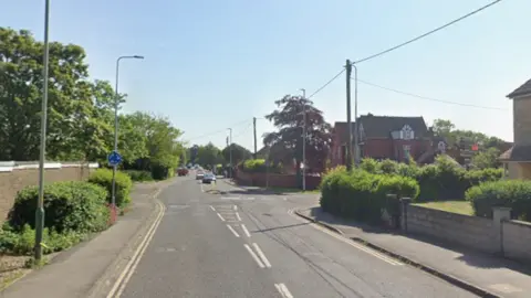 Google A leafy street, with a brick wall and trees to one side and large houses to the other. There are some cars on the road in the distance, and streetlamps and telephone wires above. 