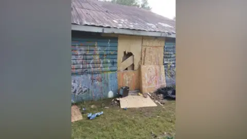 An abandoned building which is boarded up and has graffiti daubed on the walls. It has a tin roof
