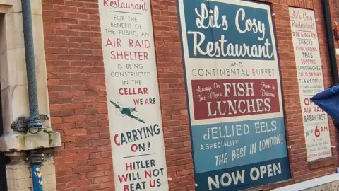 Paul Schofield Mocked up posters on a wall - one says air raid shelter being constructed in cellar - "We are carrying on - Hitler will not defeat us". Another advertises jellied eels.