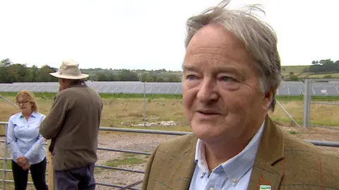 Hugh Williams of Somerset CPRE stands in front of a fence that surrounds a field of solar panels. Other campaigners are behind him.