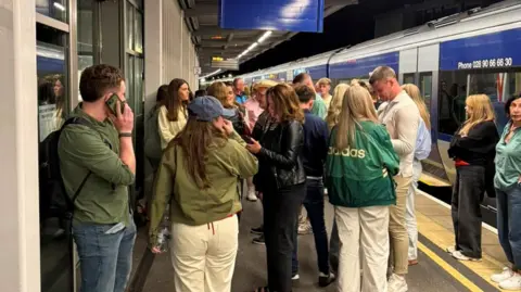 A group of passengers standing on a train platform at night. Many are looking at their mobile phones