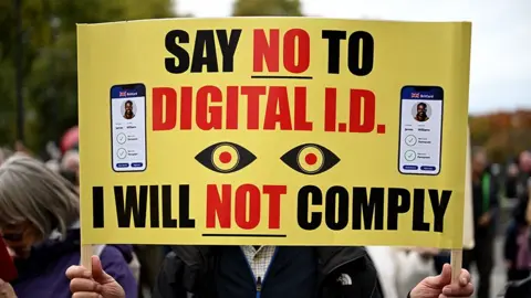A person at an anti ID card protest holding up a yellow banner which covers his face which reads "Say no to digital ID. I will not comply".