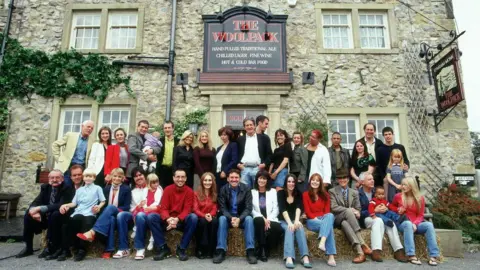A large group of adults and children pose for a photograph outside a traditional stone-built pub called The Woolpack, with several people seated on hay bales in the front row. The building has a rustic stone façade, sash windows, climbing greenery, and prominent pub signs advertising ales, wine, and food. The group stretches across the entrance and frontage of the pub, creating the appearance of a cast or community gathering in a village setting.
