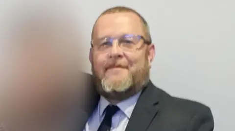 Family photo Wearing a dark suit, light-coloured shirt and dark tie, David Gamlin, with short hair and a beard, looks at the camera lens