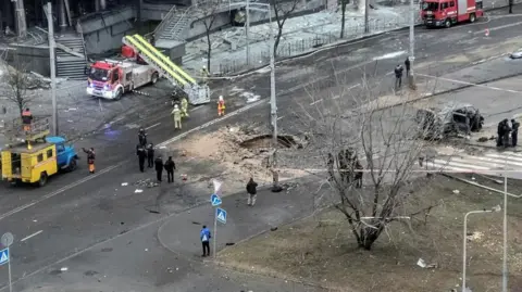 Police tape is visible as emergency vehicles and workers surround the site of a Russian missile strike in Kyiv, which left an incinerated vehicle and a hole in the road.