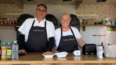 Liz Nice/BBC A man and woman stand side by side behind the counter of a van. A jacket potato with cheese and beans can be seen in front of them