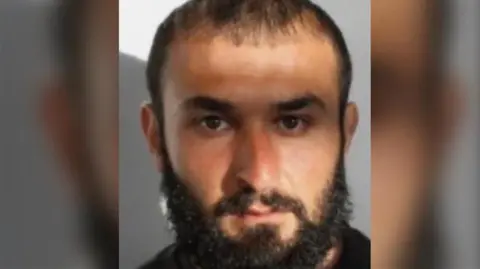 A custody image of a man with dark hair, a beard and a moustache.