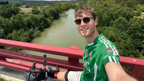 Ollie White Mr White takes a selfie from his bike, which is positioned by some red railings on a bridge over a large river. He is wearing a green sports top and dark sunglasses.