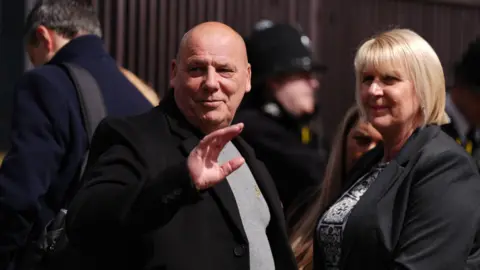Ben Whitley/PA Wire Wayne Birkett, in a grey jumper and black blazer, stands next to a woman with short blonde hair.