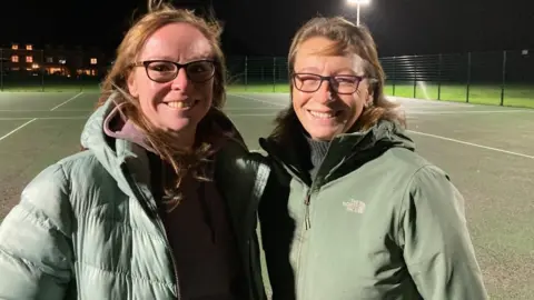 Marion Bounds and Liz Henley stand next to each other, with sports courts in the background. Liz has shoulder-length ginger hair and wears black-framed glasses and a light green puffer coat. Liz has short brown hair and wears thinner-framed black glasses and a green North Face waterproof coat.