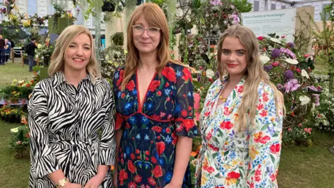 Three women stand in front of a floral display. The one on the left is wearing a zebra-print dress, while the other two have opted for floral dresses.