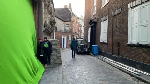 BBC A green screen is position again a wall in a city centre. Sandbags are piled high opposite a World War Two era car. 