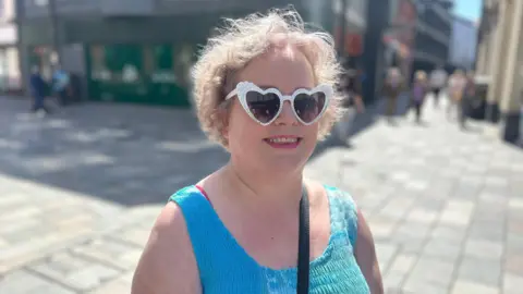 Anna-Maria Smith smiles, she wears a blue vest and white love heart-shaped sunglasses. She has short blonde hair.