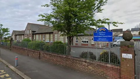 A street view of Victoria Infant and Nursery School. It is a long brick-built building, mostly single-storey with large windows and a two-storey section in the middle of the symmetrical structure. It is surrounded by a concrete playground and parking area.