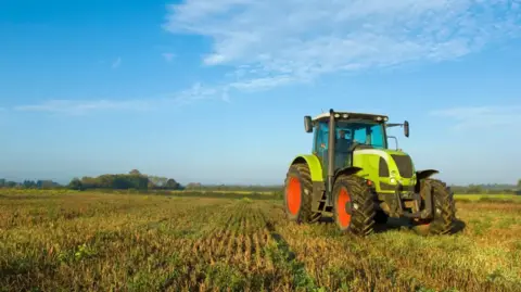 Getty Images A lime green tractor with red and black wheels in a field. There is a blue sky with high white cloud. 