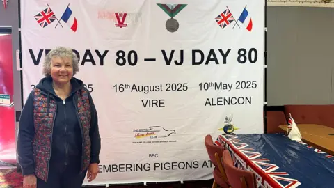 Jill Rogers stands in a room in front of a poster which reads "VE Day 80 - VJ Day 80" and lists dates of three pigeon races starting in France. She has short, grey hair and wears a gilet over a dark blue hoodie. The room also has a table with a Union Jack-decorated covering.